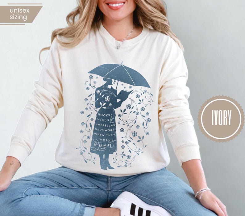 May include: Ivory-colored sweatshirt featuring a blue illustration of a person under an umbrella reading a book. The design includes floral accents and the text "Books Minds Umbrellas Only Work When They Are Open."