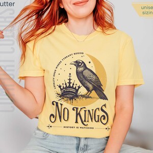 Woman wearing butter yellow Comfort Colors tees featuring a dark blue graphic of a raven, crown, and moon with No Kings – History is Watching design; Graphic is dark blue with goldenrod accents and crescent moon. unisex sizing displayed.