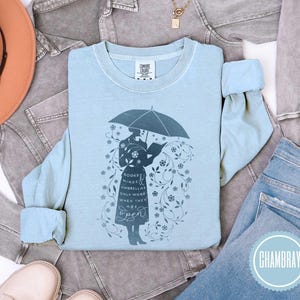 May include: Light blue long-sleeve shirt with a graphic of a person holding an umbrella and reading a book. The text on the shirt reads "Books, Minds, Umbrellas Only Work When They Are Open". The shirt is surrounded by floral designs.