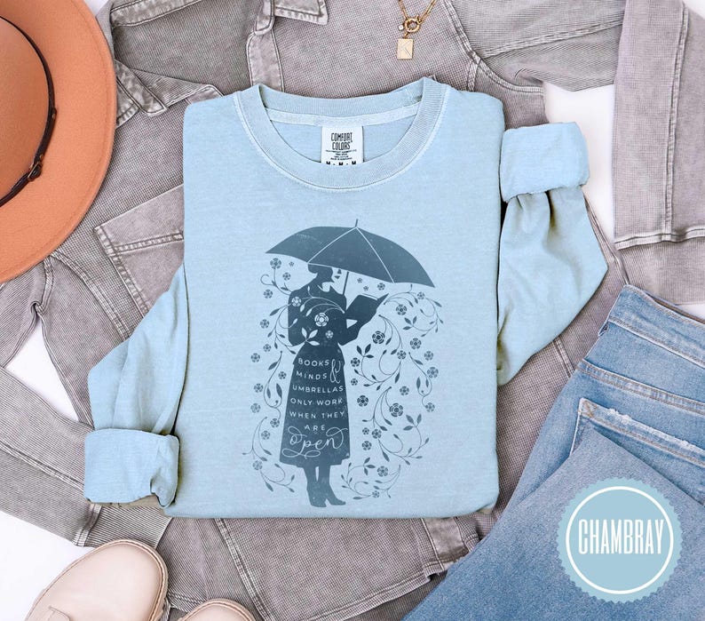 May include: Light blue long-sleeve shirt with a graphic of a person holding an umbrella and reading a book. The text on the shirt reads "Books, Minds, Umbrellas Only Work When They Are Open". The shirt is surrounded by floral designs.