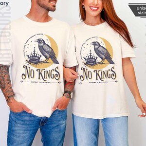 Man and woman wearing ivory Comfort Colors tees featuring a dark blue graphic of a raven, crown, and moon with No Kings – History is Watching design; Graphic is dark blue with goldenrod accents and crescent moon. unisex sizing displayed.