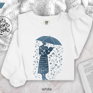 WHITE folded flat lay sweatshirt with literary umbrella design beside open book and cozy blanket, bookish home reading aesthetic
