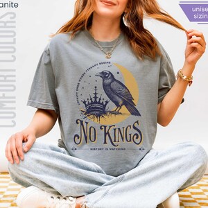Woman wearing granite (light grey) Comfort Colors tees featuring a dark blue graphic of a raven, crown, and moon with No Kings – History is Watching design; Graphic is dark blue with goldenrod accents and crescent moon. unisex sizing displayed.