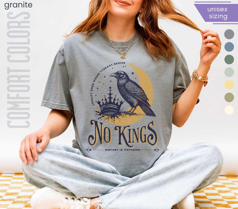 Woman wearing granite (light grey) Comfort Colors tees featuring a dark blue graphic of a raven, crown, and moon with No Kings – History is Watching design; Graphic is dark blue with goldenrod accents and crescent moon. unisex sizing displayed.
