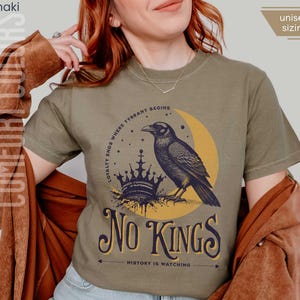 Woman wearing khaki Comfort Colors tees featuring a dark blue graphic of a raven, crown, and moon with No Kings – History is Watching design; Graphic is dark blue with goldenrod accents and crescent moon. unisex sizing displayed.