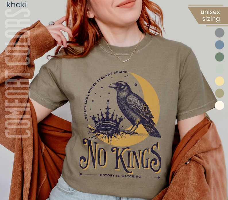 Woman wearing khaki Comfort Colors tees featuring a dark blue graphic of a raven, crown, and moon with No Kings – History is Watching design; Graphic is dark blue with goldenrod accents and crescent moon. unisex sizing displayed.