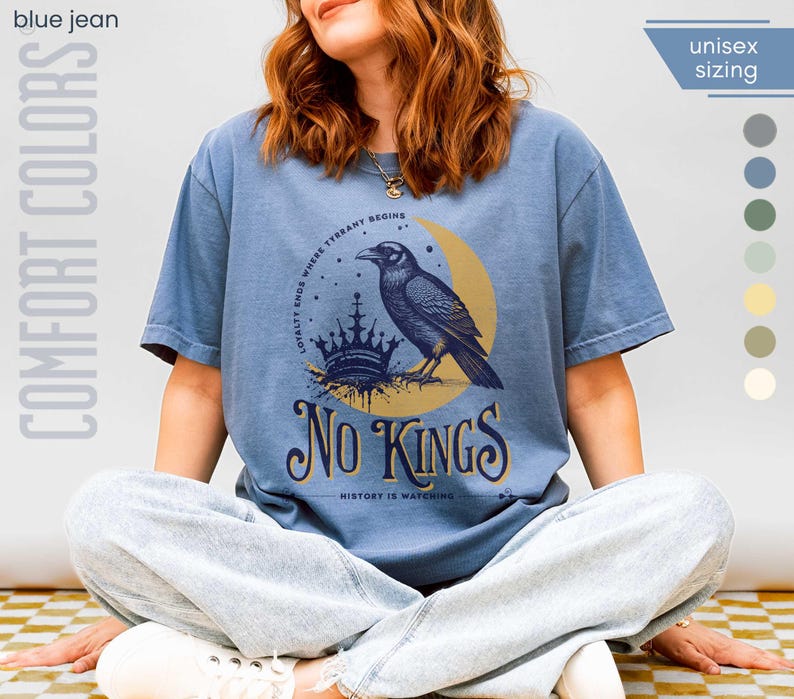 Woman wearing blue jean (muted medium slate blue) Comfort Colors tees featuring a dark blue graphic of a raven, crown, and moon with No Kings – History is Watching design; Graphic is dark blue with goldenrod accents and crescent moon. unisex sizing displayed.