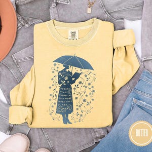 May include: A yellow long-sleeve shirt with a blue graphic of a person under an umbrella reading a book. The shirt has the text "Books, Minds, Umbrellas Only Work When They Are Open". The shirt is surrounded by denim and a hat.