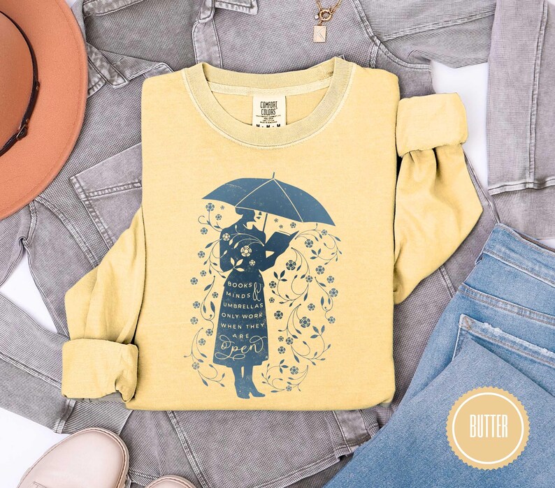 May include: A yellow long-sleeve shirt with a blue graphic of a person under an umbrella reading a book. The shirt has the text "Books, Minds, Umbrellas Only Work When They Are Open". The shirt is surrounded by denim and a hat.