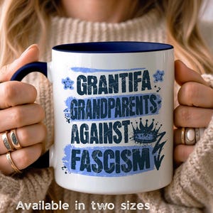 May include: White ceramic mug with a navy blue interior and handle. The mug features the text "GRANTIFA GRANDPARENTS AGAINST FASCISM" in blue, with a crown and star design. The mug is held by a person.