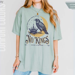 Woman wearing bay (light muted green) Comfort Colors tees featuring a dark blue graphic of a raven, crown, and moon with No Kings – History is Watching design; Graphic is dark blue with goldenrod accents and crescent moon. unisex sizing displayed.
