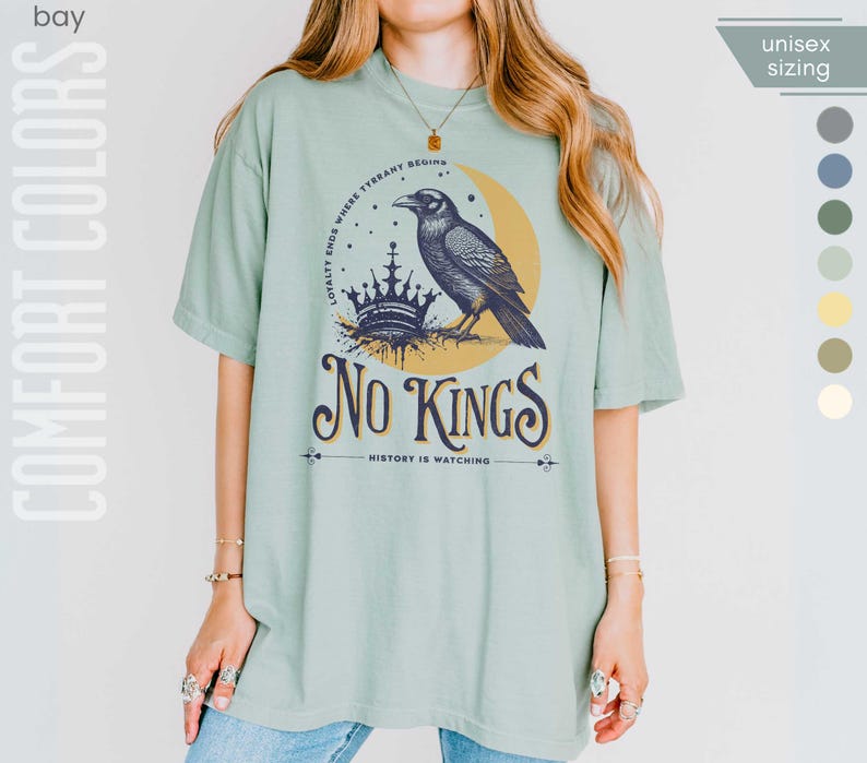 Woman wearing bay (light muted green) Comfort Colors tees featuring a dark blue graphic of a raven, crown, and moon with No Kings – History is Watching design; Graphic is dark blue with goldenrod accents and crescent moon. unisex sizing displayed.