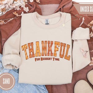May include: A sand-colored sweatshirt with the text "THANKFUL FOR DESSERT TIME" in orange and brown block letters. The sweatshirt has a crew neck and long sleeves. The words "UNISEX SIZING" are in the upper right corner.