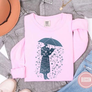 May include: A light pink long-sleeved shirt with a graphic of a person under an umbrella reading a book, surrounded by floral elements. The text on the shirt reads "Books, Minds, Umbrellas Only Work When They Are Open."