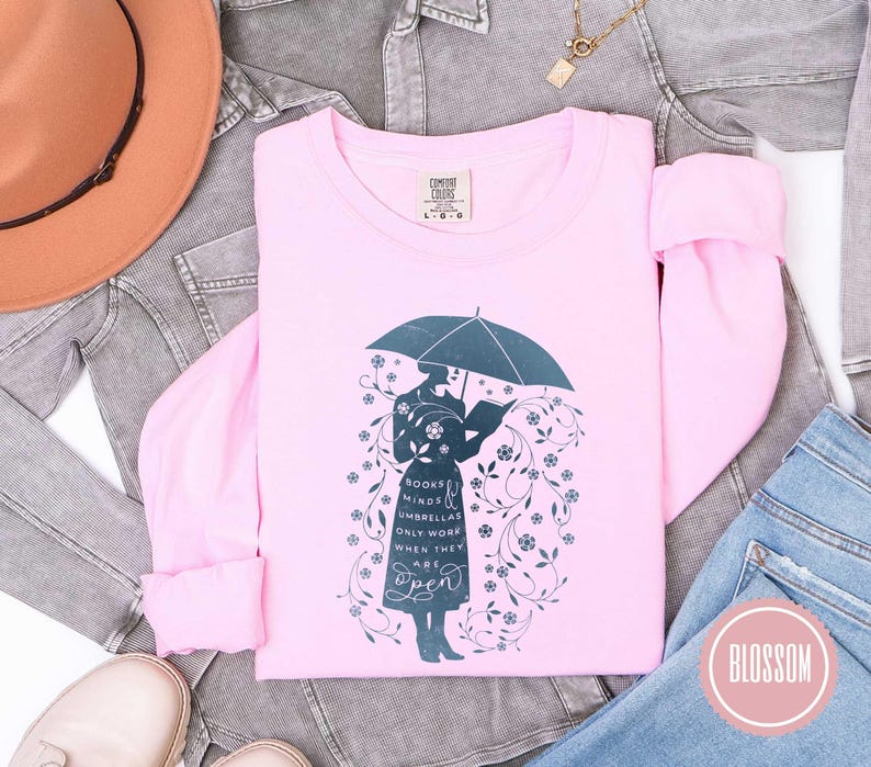 May include: A light pink long-sleeved shirt with a graphic of a person under an umbrella reading a book, surrounded by floral elements. The text on the shirt reads "Books, Minds, Umbrellas Only Work When They Are Open."