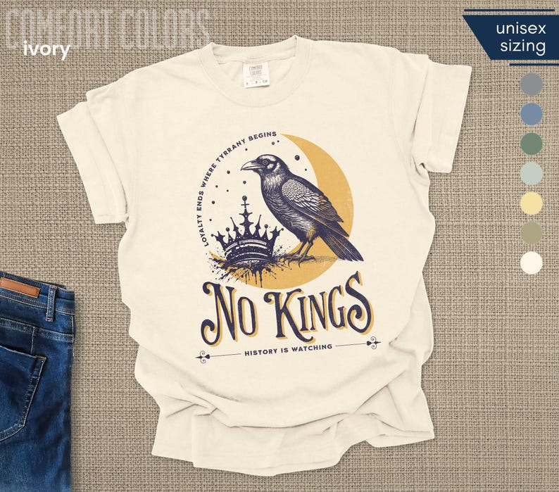 Flat lay of ivory Comfort Colors tee with a dark blue raven perched on crown and a goldenrod colored crescent moon, reading No Kings – History is Watching, on a textured background with jeans.