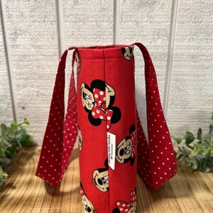 May include: A red tote bag featuring a repeating pattern of Minnie Mouse. The bag has red polka-dot handles and a small white tag. The bag is made of fabric and is designed to carry items.