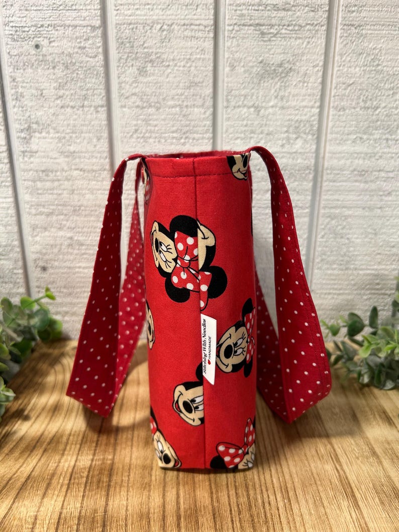 May include: A red tote bag featuring a repeating pattern of Minnie Mouse. The bag has red polka-dot handles and a small white tag. The bag is made of fabric and is designed to carry items.