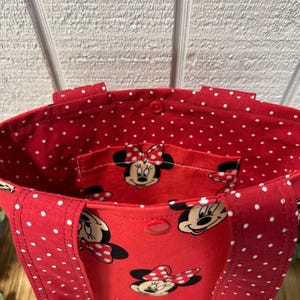 May include: Red tote bag with white polka dots and a Minnie Mouse print. The bag has a red interior with a small pocket featuring Minnie Mouse. The handles and exterior are red with white polka dots.