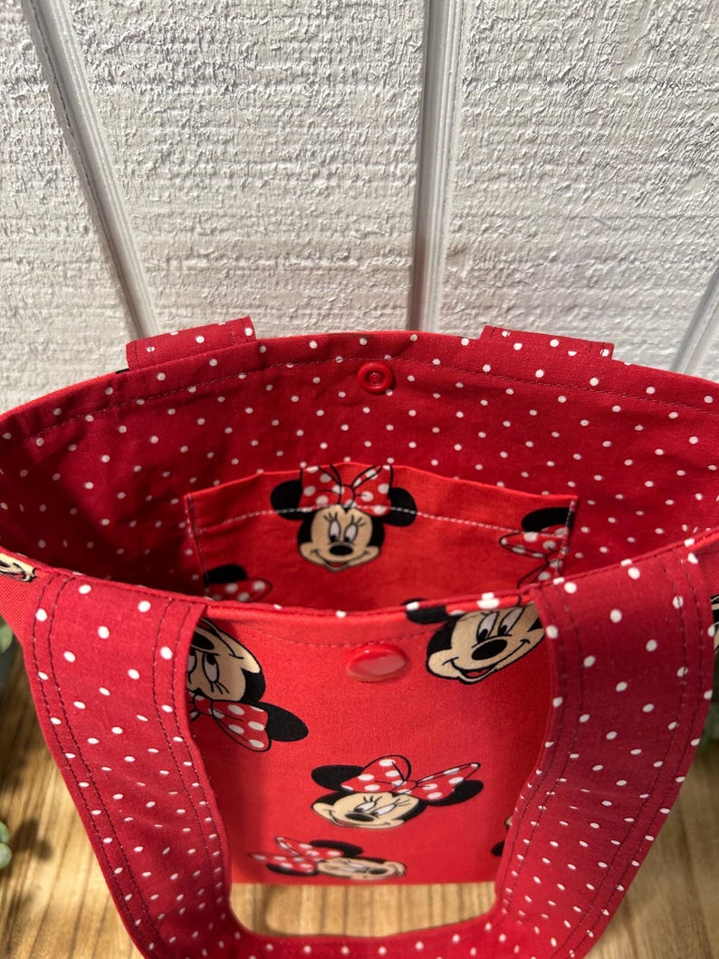 May include: Red tote bag with white polka dots and a Minnie Mouse print. The bag has a red interior with a small pocket featuring Minnie Mouse. The handles and exterior are red with white polka dots.