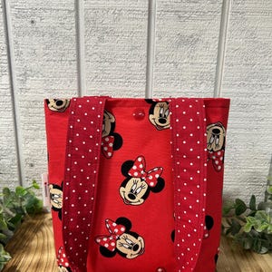 May include: Red tote bag featuring a repeating pattern of Minnie Mouse's face with a red bow. The bag has red handles with white polka dots. The background is a light wood panel.