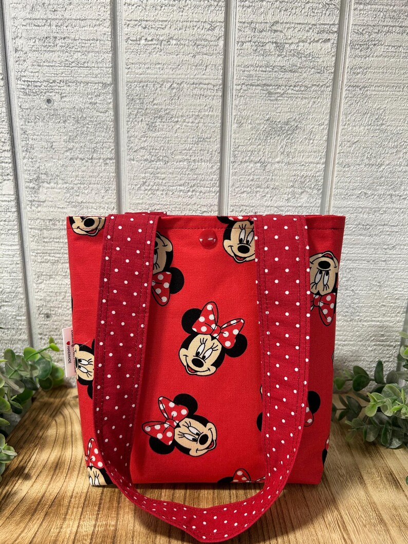 May include: Red tote bag featuring a repeating pattern of Minnie Mouse's face with a red bow. The bag has red handles with white polka dots. The background is a light wood panel.