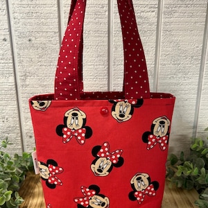 May include: A red tote bag featuring a pattern of Minnie Mouse faces with red and white polka dot bows. The bag has red handles with white polka dots and a red interior. A red button secures the top.