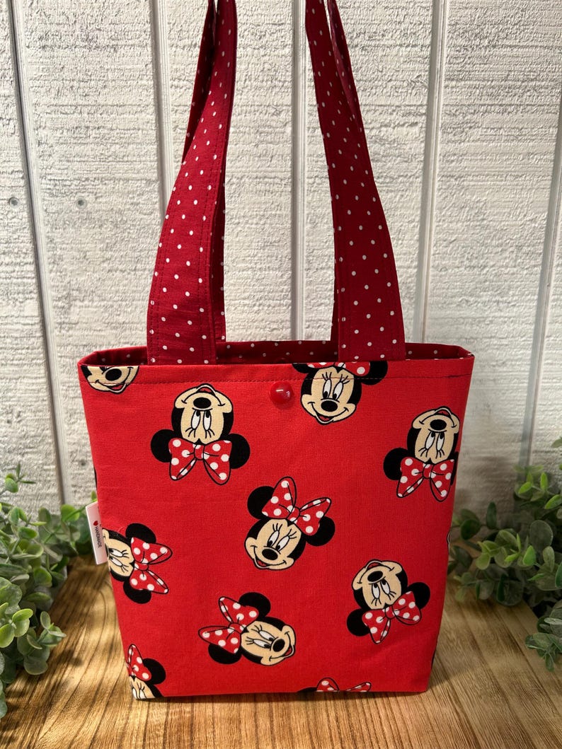 May include: A red tote bag featuring a pattern of Minnie Mouse faces with red and white polka dot bows. The bag has red handles with white polka dots and a red interior. A red button secures the top.