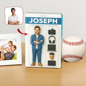 Może przedstawiać: Spersonalizowana figurka akcji o imieniu JOSEPH, w dżinsowej kurtce i dżinsach, obok piłki do baseballu i oprawionego zdjęcia. Figurka zawiera miniaturowe akcesoria: aparat fotograficzny, słuchawki, laptop, kubek do kawy i tablet. Opakowanie ma niebieski baner z imieniem JOSEPH.