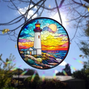 May include: A stained glass suncatcher depicting a lighthouse on a rocky shore with a sunset sky and ocean waves. The suncatcher is round and has a metal chain for hanging.