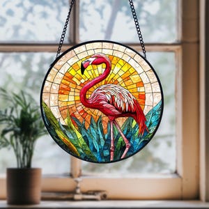 Puede incluir: Una ventana de vidrio emplomado colgada con un flamenco rosa de pie en un lecho de plantas azules y verdes. El fondo es una puesta de sol amarilla y naranja.