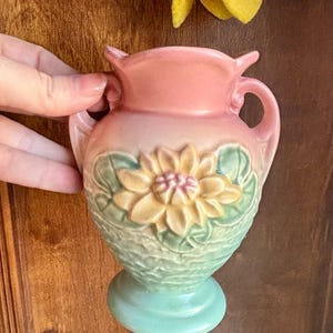 May include: A small, vintage ceramic vase in shades of pink, green, and yellow. The vase features a floral design on the front and two handles. The base is a light teal color. The vase is held by a hand.