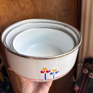 May include: Set of three white enamel mixing bowls with silver rims. Each bowl has a floral design in red, yellow, and blue. A red stripe encircles the bowls near the bottom. The bowls are nested together.