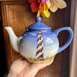 May include: A ceramic teapot with a blue lid and handle, and a white spout. The body of the teapot features a lighthouse design with brown and white stripes, set against a blue sky and green grass.