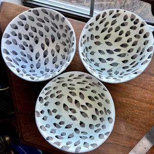 May include: Three white ceramic bowls with a black and white leaf pattern. The bowls are on a wooden surface.