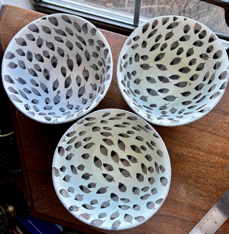 May include: Three white ceramic bowls with a black and white leaf pattern. The bowls are on a wooden surface.