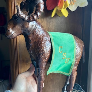 May include: A brown ram sculpture with large, curved horns and a green banner embroidered with "CSU". The ram has a textured surface and is held in front of a wooden cabinet. A colorful felt flower arrangement is in the background.