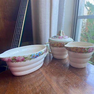 May include: A vintage white ceramic creamer, sugar bowl, and gravy boat set with a floral pattern in shades of pink, blue, green, and yellow. The set has a white background with a green border and a floral design around the top edge.