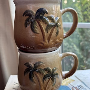 May include: Two stacked ceramic mugs with a tan and brown gradient glaze. Each mug features a raised, embossed design of two palm trees and small birds. The top mug has the word Hawaii written in cursive script at the base of the palm trees.