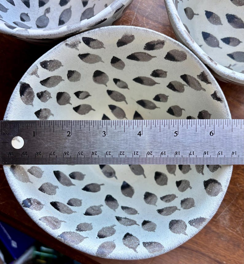 May include: A white ceramic bowl with a black leaf pattern. The bowl is 6 inches in diameter and is sitting on a wooden surface.
