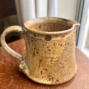 May include: A small, speckled brown and cream ceramic creamer with a handle. The creamer has a rustic, handmade look.