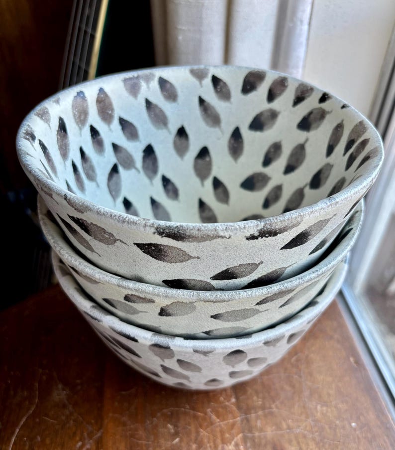 May include: Three white ceramic bowls with a black leaf pattern. The bowls are stacked on top of each other.