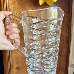 May include: A clear glass pitcher featuring a prominent geometric diamond-cut pattern across its exterior surface. The vessel includes a sturdy integrated handle on the side and a wide pouring spout at the top rim, showcasing a faceted design.