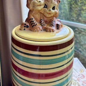 May include: A ceramic canister with a lid featuring two sculpted tiger cubs. The canister body has a cream-colored base with horizontal stripes in dark brown, teal, and deep red. The tiger cubs are orange with black stripes and painted facial features.