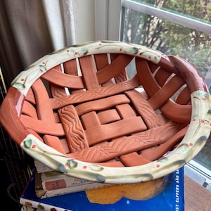 May include: A handmade, oval-shaped ceramic basket with a woven design in shades of terracotta. The rim is decorated with a cream-colored band featuring green leaves and red berries. The basket is sitting on top of a stack of books.