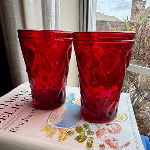 May include: Two ruby red drinking glasses with a textured pattern. The glasses are cylindrical and sit on a cookbook. The book's title is "Simply Delicious" by Eva Jean S. The glasses are near a window.