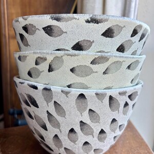 May include: A set of three white ceramic bowls with a black leaf pattern. The bowls are stacked on top of each other.