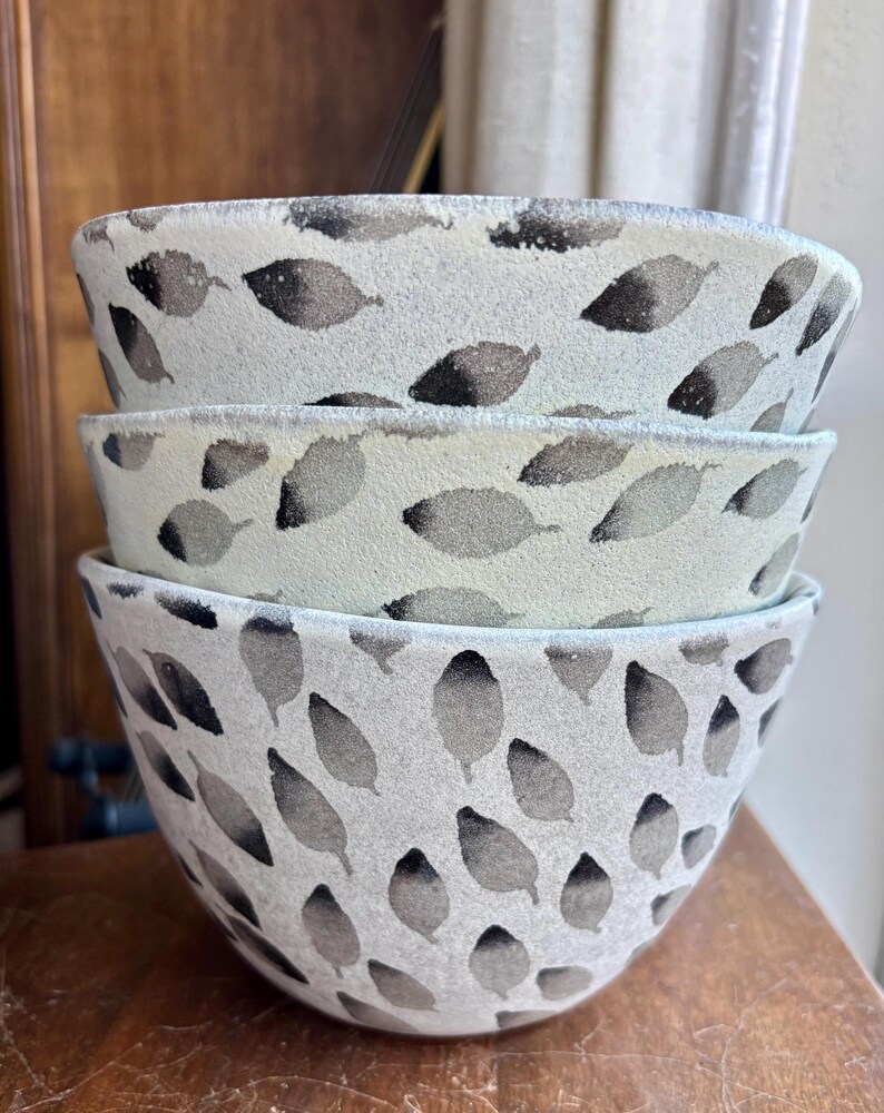 May include: A set of three white ceramic bowls with a black leaf pattern. The bowls are stacked on top of each other.