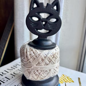 May include: A black cat-shaped yarn holder with a smiling face, holding a beige ball of yarn. The yarn holder is made of metal and sits on a book. The yarn ball is approximately 4 inches in diameter.