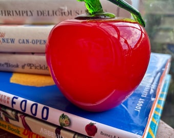 May include: A vibrant red glass apple with a green stem sits atop a stack of books. The apple has a glossy finish. The books include titles like "Shrimply Delicious" and "Dictionary in French".